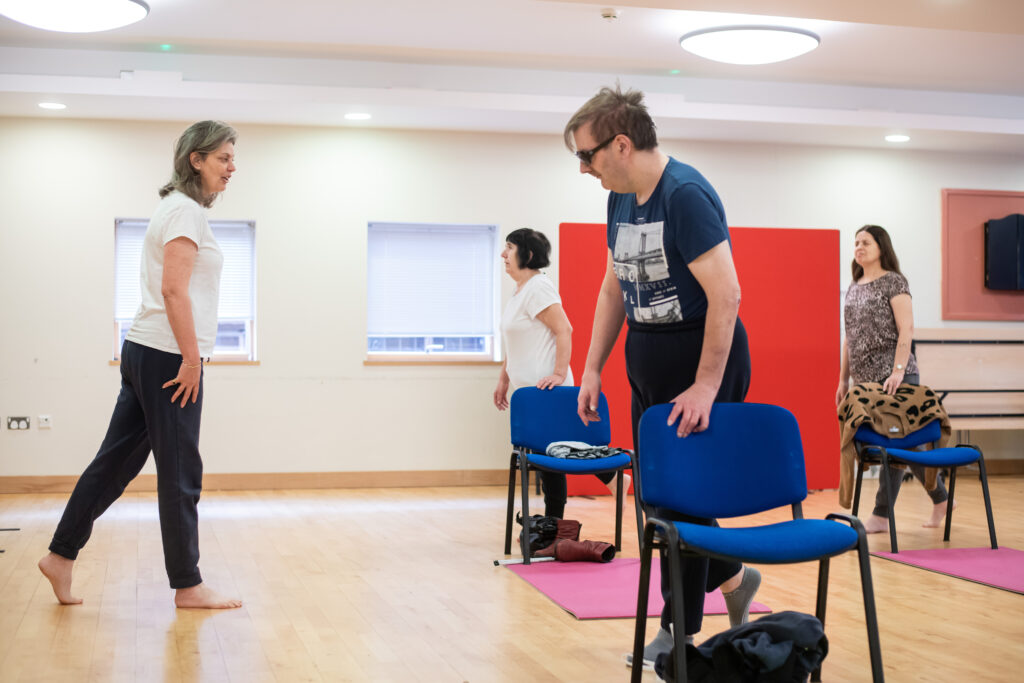 Image: A session of the weekly yoga class in the Norwich Hub. Three participants stand on one foot behind chairs and yoga mats while the instructor watches from the front of the class